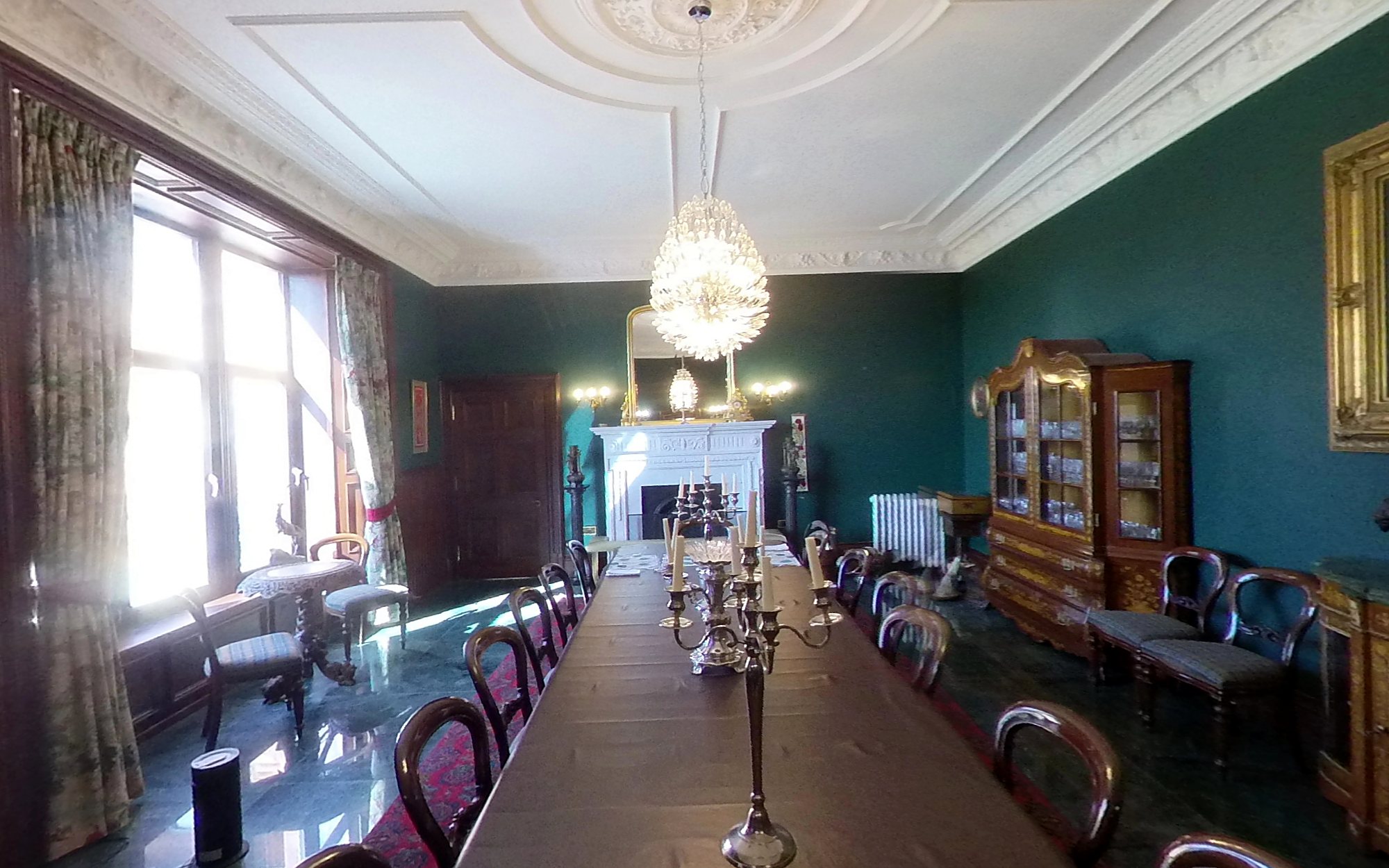 The same dining room in April 2025: green walls, white plaster ceiling, chandelier and long table, with sun through the garden window
