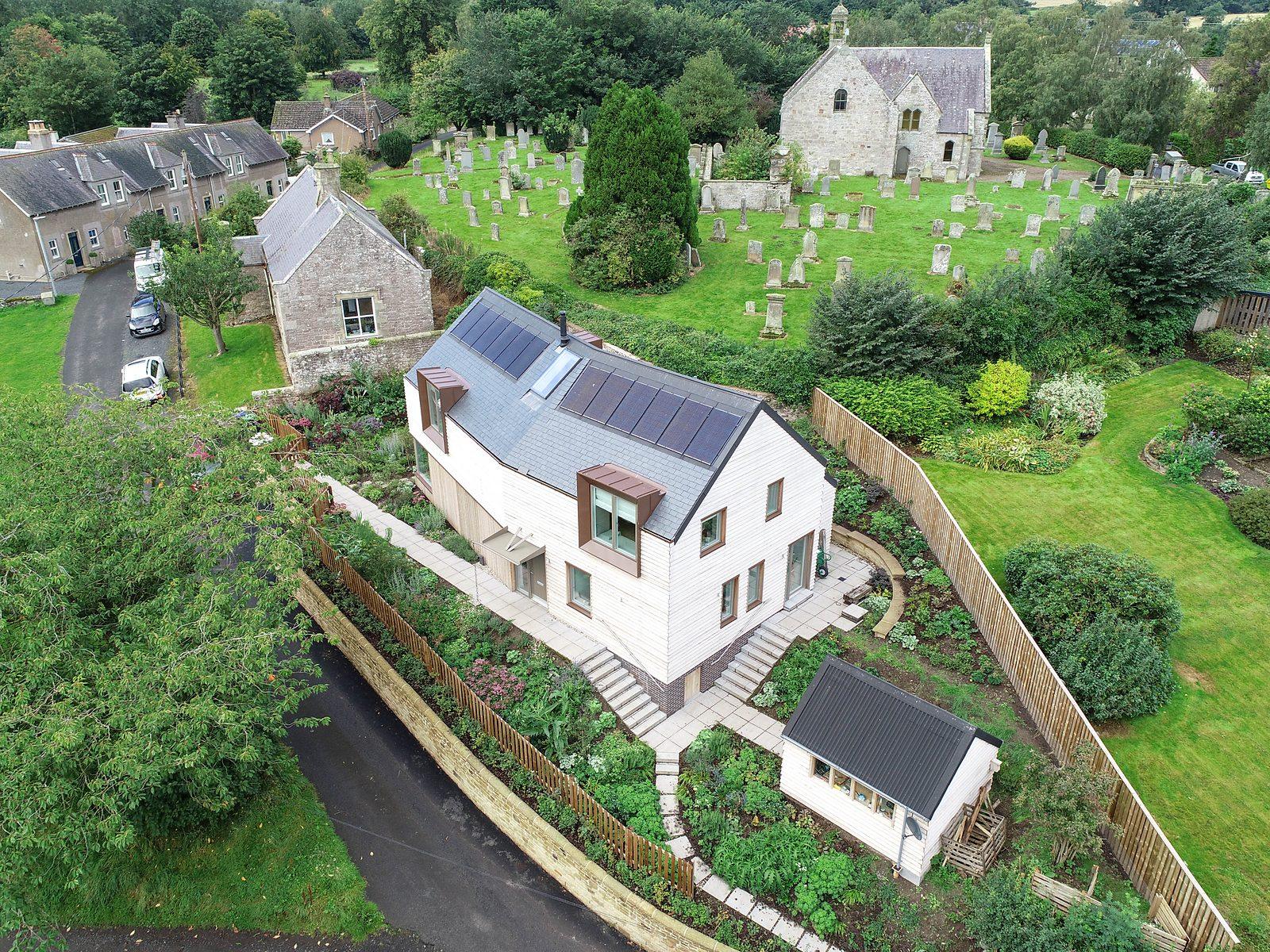 Pale new house with a grey roof on a sloping garden beside a village churchyard